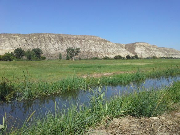 Across Highway 52 to the north, working ranches, community irrigation canals, and distantly recognizable sandstone cliffs and bluffs lie in close proximity to the directionally drilled and potentially fracked Smoke Ranch natural gas well, within a river confluence and wetlands floodplain.