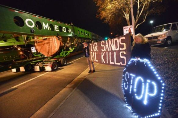 On Monday, December 2, 2013, light brigade action team protesters Rod Lyman and Kathy Leathers of Bellingham, Washington, hold up signs, as a megaload slowly passes by on Highway 395 in Hermiston, Oregon.  The transport rig carries a 450-ton piece of equipment bound for a tar sands development site in western Canada (Associated Press/E.J. Harris photo).